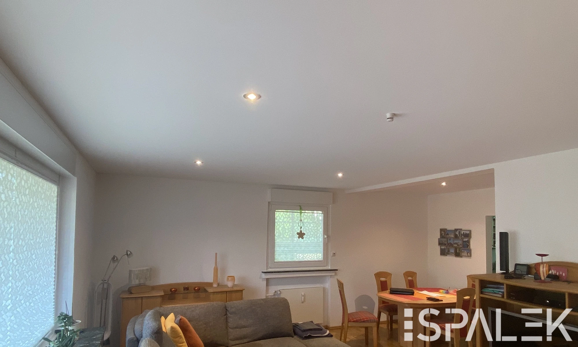 Wohn- und Essbereich mit grauem Sofa, bunten Kissen, Holztisch, Stühlen, Schränken und Fensterjalousie. Spanndecke mit Beleuchtung.