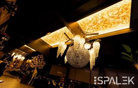 Elegantes Restaurant mit großer runder Glaslampe, Kristallleuchten und floral gemusterter, hinterleuchteter Spanndecke.