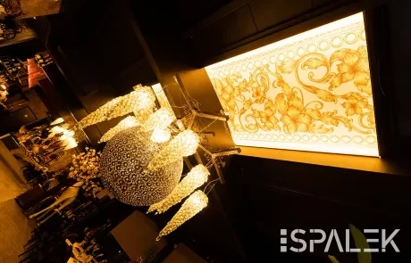 Ein Kristalllüster mit verzierten Glasdetails hängt an einer Spanndecke neben einer leuchtenden Paneel mit floralen Mustern.