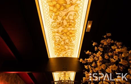 Ein warm beleuchteter Innenraum mit einer kunstvoll verzierten Spanndecke mit goldenen floralen Mustern, Kronleuchtern und Blumenvasen.