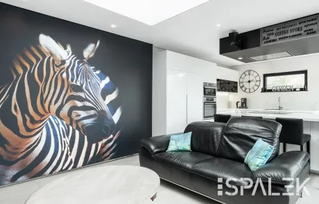 Wohnzimmer Modernes Wohnzimmer mit großem Zebramotiv, schwarzem Ledersofa, blauem Kissen, rundem Holztisch und weißer Küche unter Spanndecke.