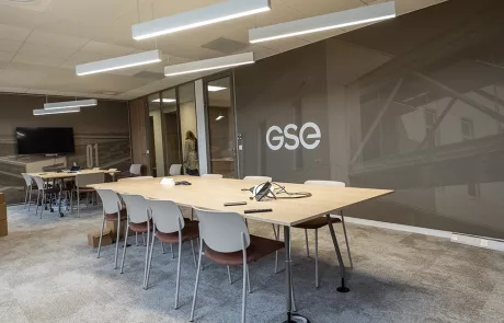 Akkustik Beispiel_1 Moderner Konferenzraum mit langem Holztisch, weißen Stühlen, Deckenbeleuchtung, TV mit "GSE"-Logo, Glastüren und Teppichboden.
