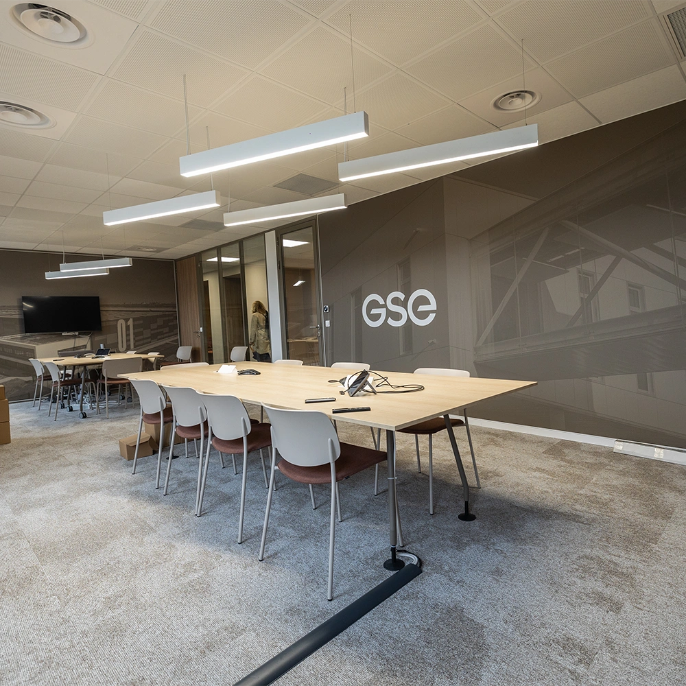 Moderner Konferenzraum mit langem Holztisch, weißen Stühlen, Deckenbeleuchtung, TV mit "GSE"-Logo, Glastüren und Teppichboden.