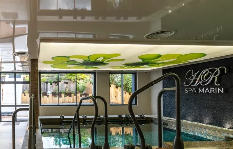 Akkustik Beispiel_2 Innenbereich eines Spa-Pools mit Stufen ins Wasser, grünen Blättern an der Decke, großen Fenstern und HOR SPA MARIN Schild an der Wand.