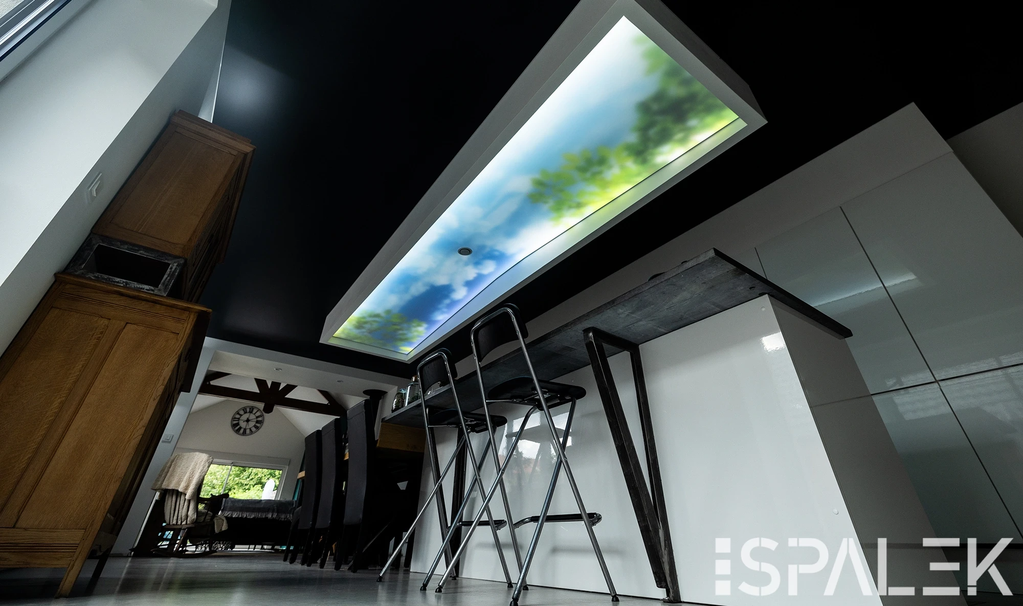Moderne Küche mit weißen Schränken, hohem Holzschrank, schwarzer Kochinsel und rechteckigem Lichtpanel in Himmel-Optik.