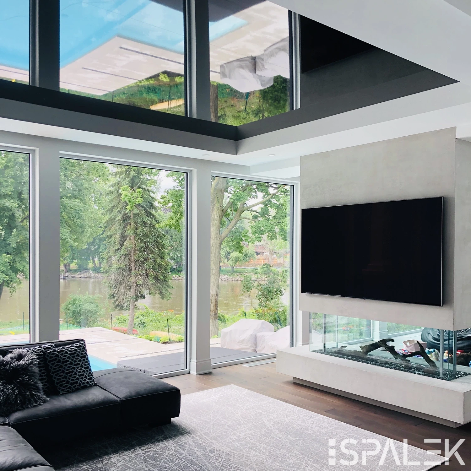 Modernes Wohnzimmer mit Akustikdecke, bodentiefen Fenstern mit Blick auf Bäume und Fluss, schwarzem Sofa und Glas-Kamin.