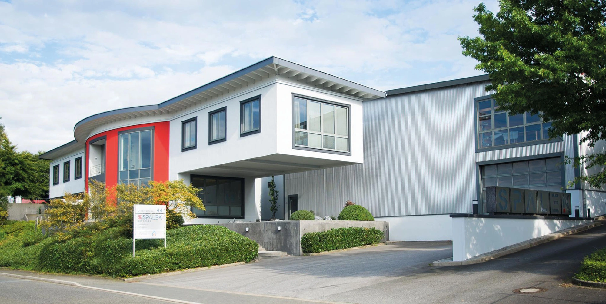 Modernes Gewerbegebäude mit weißen und roten Fassaden, großen Fenstern, begrünten Außenanlagen und Zufahrt zum Eingang.