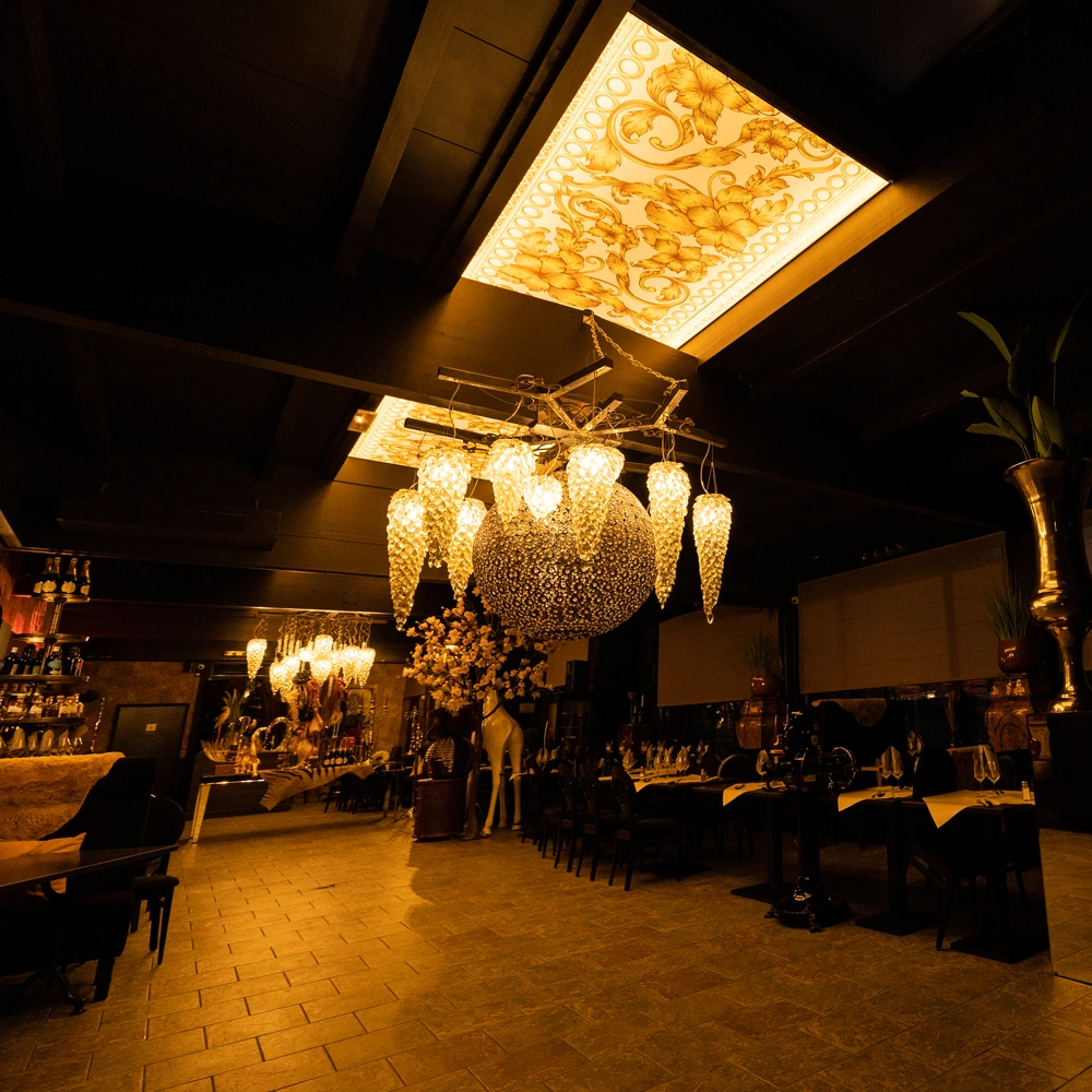 Ein stilvolles, schummrig beleuchtetes Restaurant mit goldenen Kronleuchtern, Akustikdecke, verzierten Deckenpanelen und gedeckten Tischen.