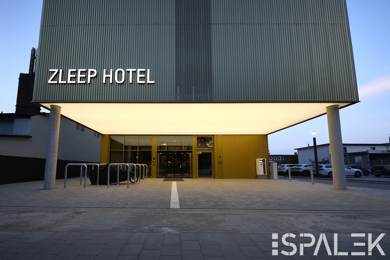 Der Eingang des Zleep Hotel Hamburg zeigt eine moderne Fassade mit großem Leuchtschild, Glastüren, Fahrradständern und breitem Weg bei Dämmerung.