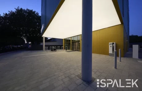 Modernes Gebäude mit gelber Metallfassade und beleuchtetem Vordach am Abend; davor Fahrradständer, Bäume und ein Geldautomat.