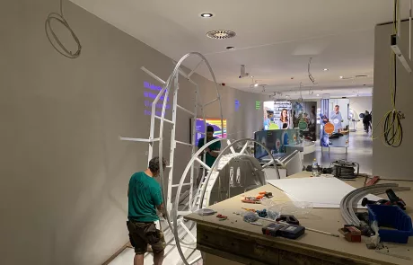 Eine Person im grünen Shirt montiert eine große Metallkonstruktion in einem hellen, im Bau befindlichen Raum des Museums Sensoria Holzminden; Werkzeuge und Materialien liegen verstreut, Ausstellungsstücke sind im Hintergrund sichtbar.
