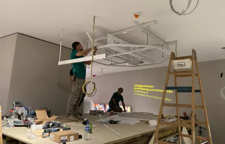 Zwei Arbeiter montieren auf einer Hebebühne eine Metallunterkonstruktion an der Decke eines Rohbaus im Museum Sensoria.