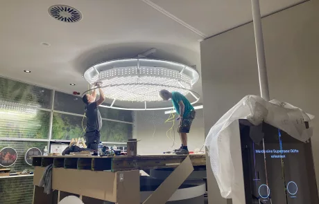 Zwei Personen montieren auf einer Hebebühne ein großes rundes LED-Deckenlicht im Rohbau des Museum Sensoria, Holzminden.
