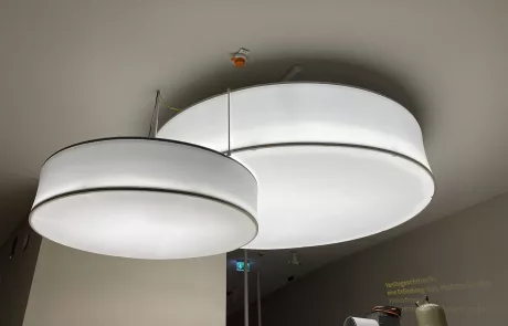 Große, moderne, runde Deckenleuchten mit weißen Diffusoren hängen an einer weißen Decke im Museum Sensoria in Holzminden. Darunter sind Baumaterialien und Geräte zu sehen.