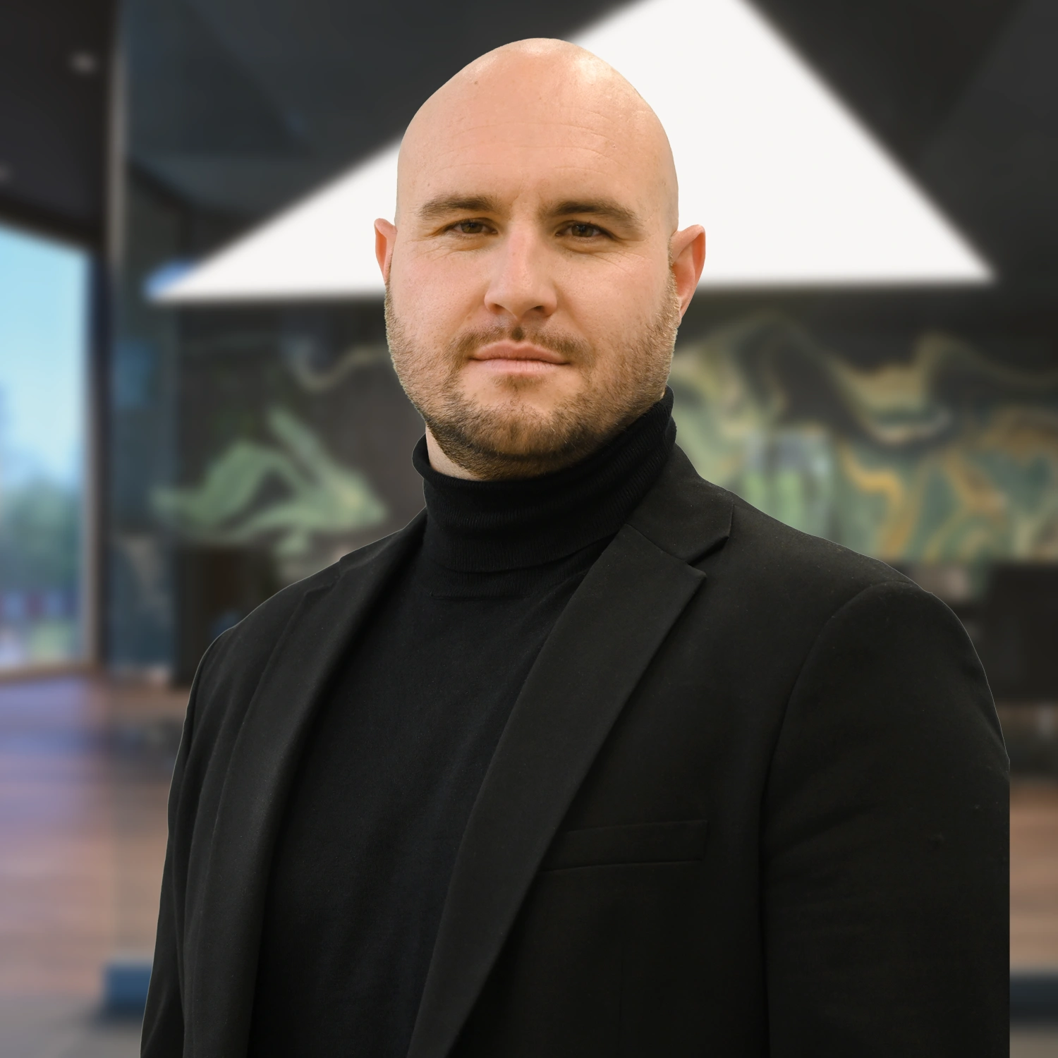 Ein Mann mit rasiertem Kopf und Bart trägt schwarzen Rollkragenpullover und Blazer vor unscharfem, modernem Hintergrund.