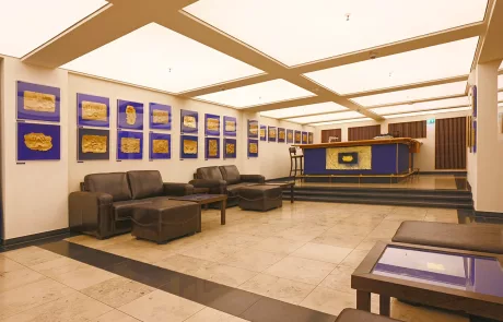 Lichtburg1 Großzügige Lobby mit braunen Ledersofas, Tischen, beigem Fliesenboden und blau gerahmten goldenen Wandbildern. Rezeption mit blauem Frontpanel unter heller Deckenbeleuchtung.