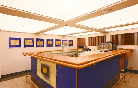 Lichtburg2 Moderne Kaffeebar mit Holztresen und blauen Akzenten, großer Espressomaschine, heller Beleuchtung sowie abstrakter Wandkunst.
