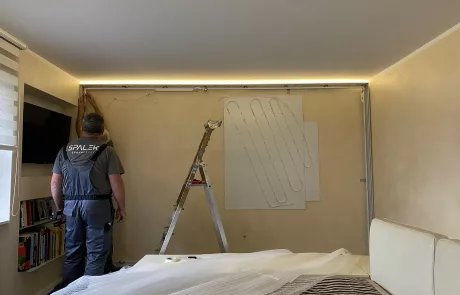 Montage1 Eine Person in Arbeitskleidung steht neben einem Leiter und arbeitet an der Wand; Möbel sind abgedeckt, warmes Licht im Raum.