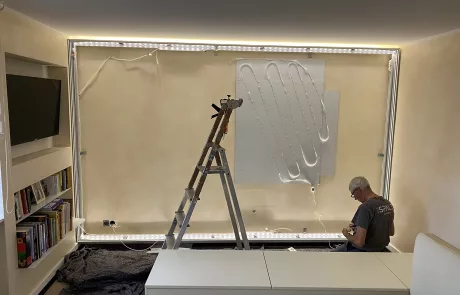 Montage2 Ein Mann arbeitet am Boden an Elektrokabeln entlang einer hellen Wand mit LED-Streifen; daneben Leiter und Bücherregale.