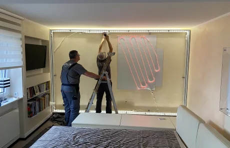Montage3 Zwei Personen montieren ein Heizpanel mit leuchtend roten Heizspiralen an einer Wand in einem modernen, aufgeräumten Raum.