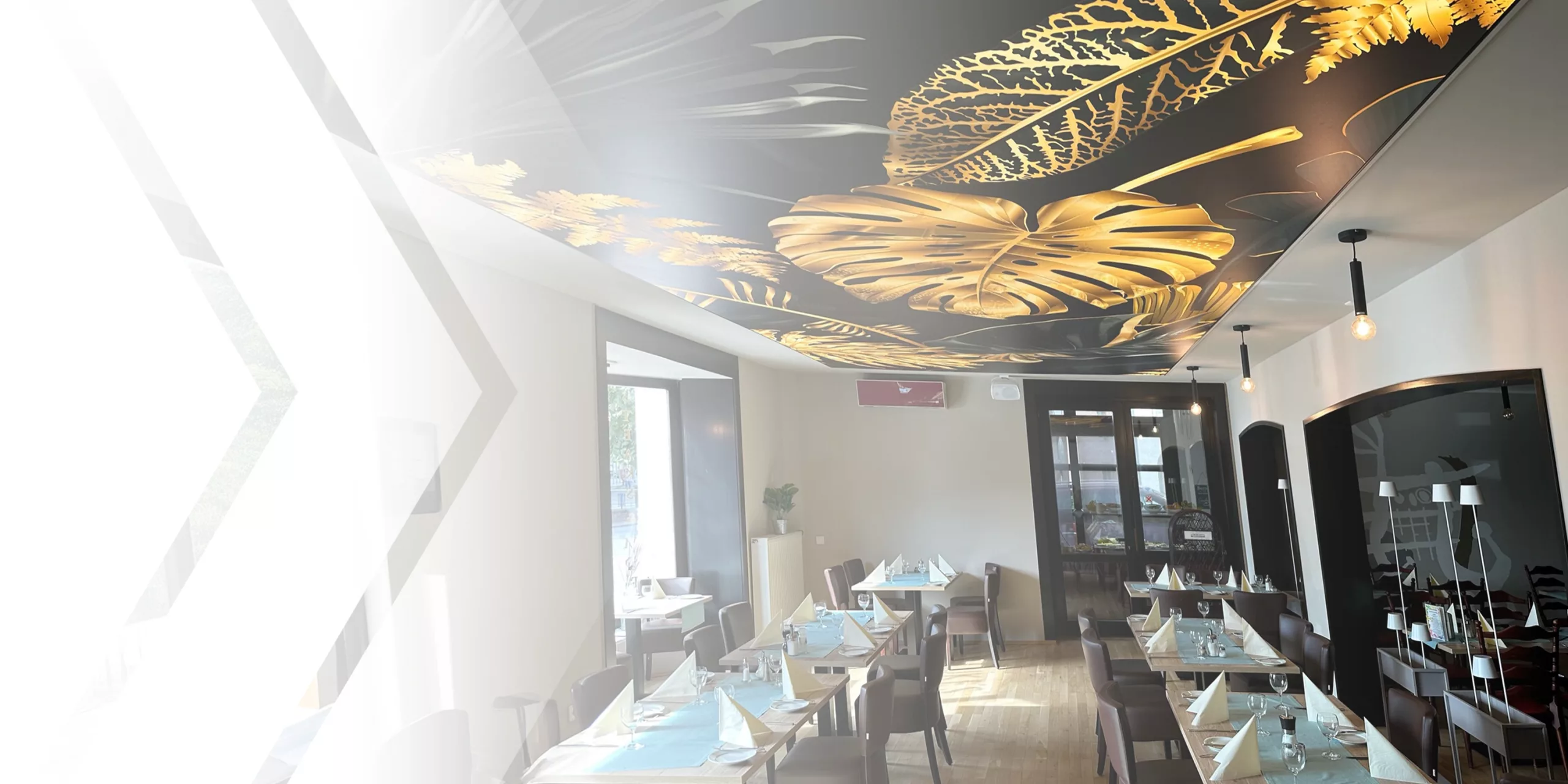 Modernes Restaurant mit gepflegten gedeckten Tischen, großen Fenstern und einer Decke mit goldenen Blattmustern. Warmes Licht schafft Gemütlichkeit.