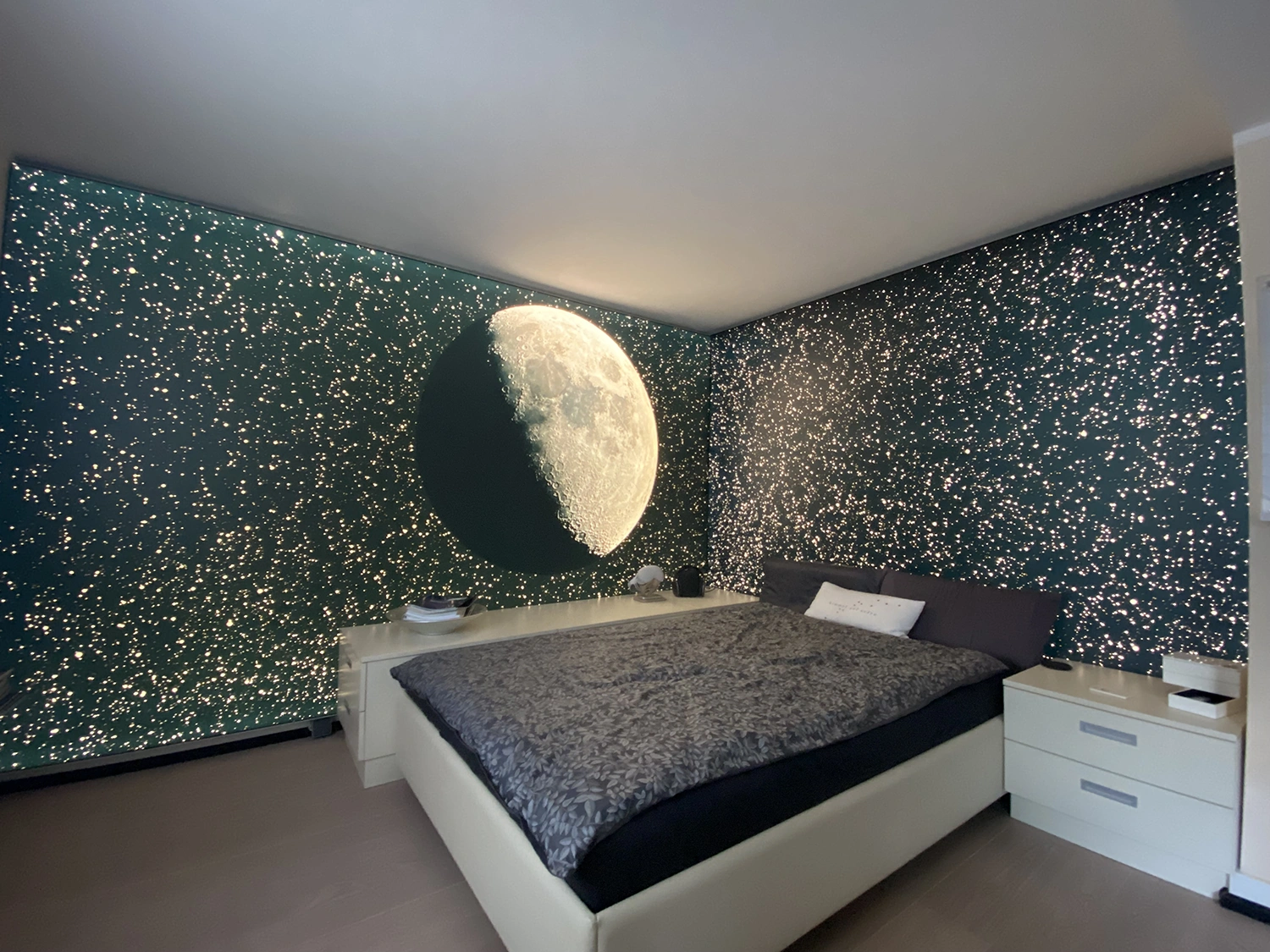 Schlafzkomplett Modernes Schlafzimmer mit dunkelgrünen Wänden, kleinen weißen Lichtern und großem Mondmotiv über einem Bett mit grauer Bettwäsche.