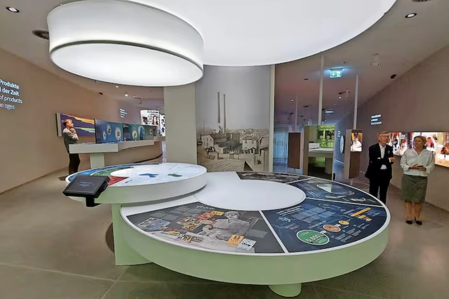Moderne Museumsausstellung in Holzminden mit runden interaktiven Displays, zwei Personen und heller Beleuchtung durch Deckenleuchten.