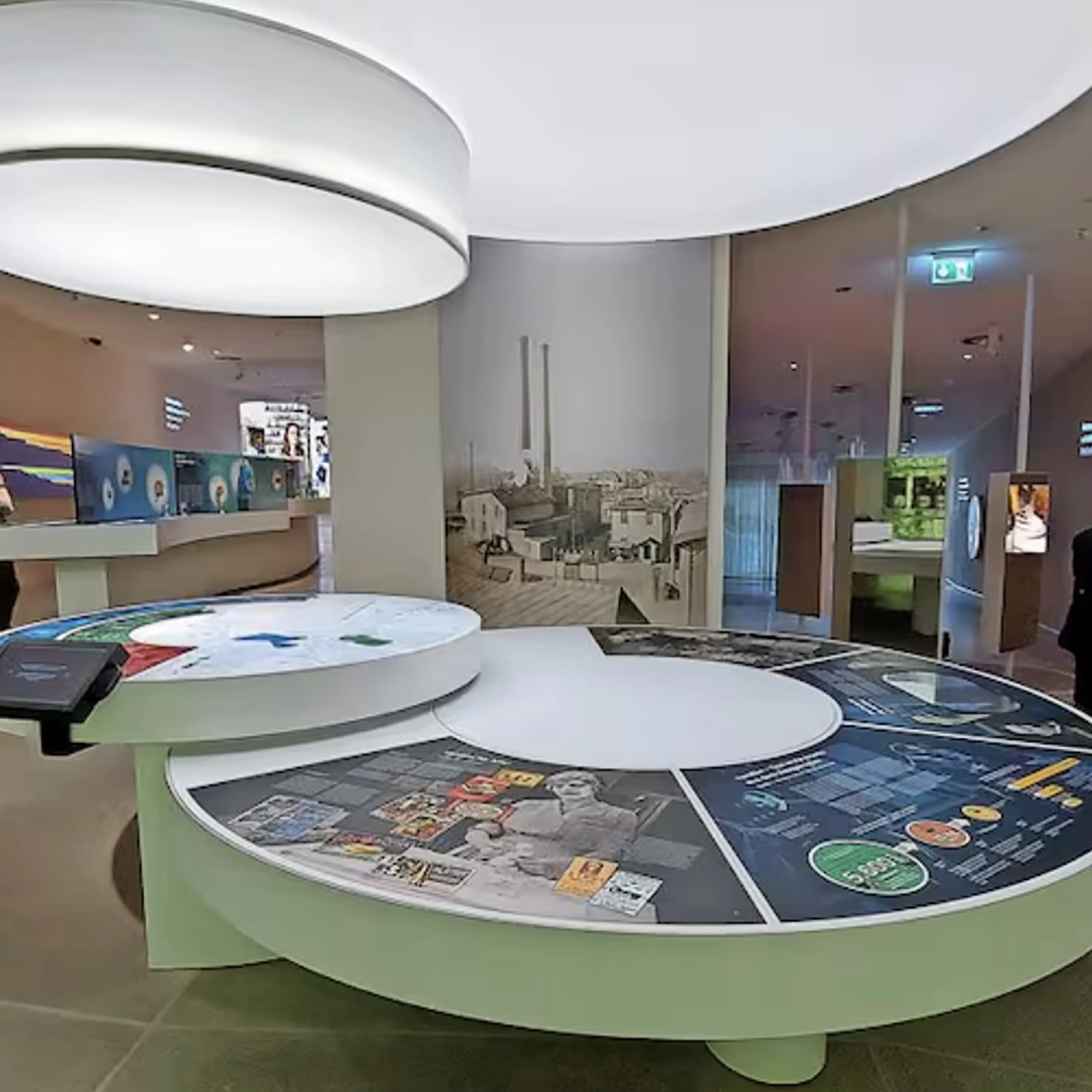 Im Museum Sensoria in Holzminden stehen große runde Medientische, historische Fotos, Infotafeln und ein Fabrikfoto an der gebogenen Wand.