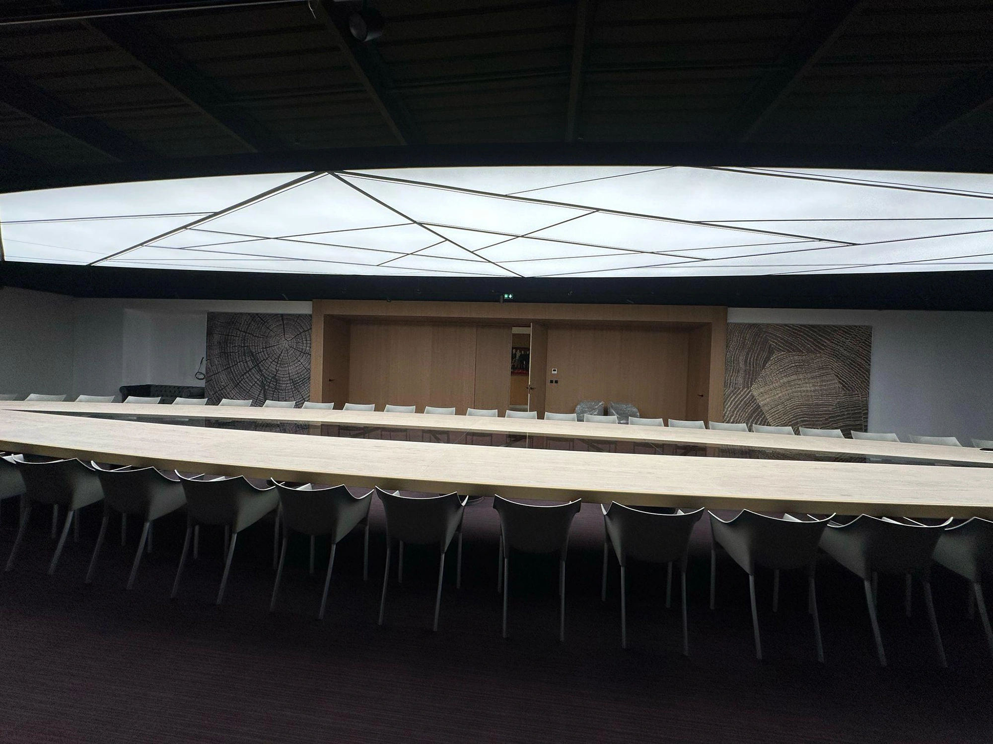 17 m Leuchte 3 Moderner Konferenzraum mit langem Holztisch, weißen Stühlen, geometrischer Deckenbeleuchtung und holzgemusterter Wandkunst.