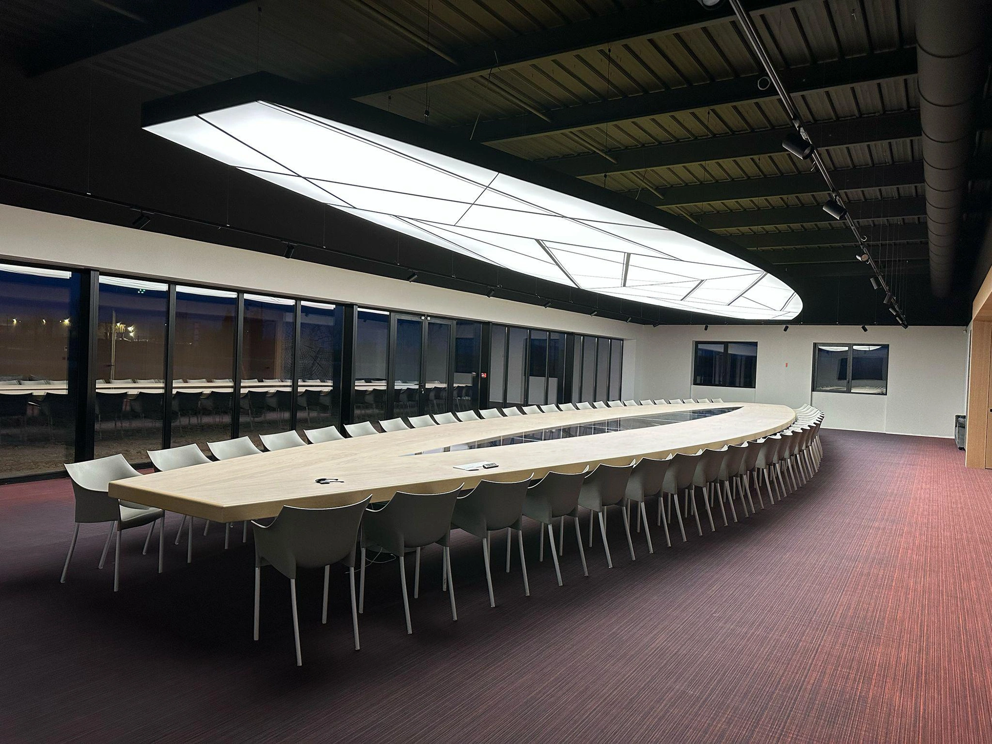 17 m Leuchte 5 Großer, moderner Konferenzraum mit langem, ovalem Tisch, weißen Stühlen, bodentiefen Fenstern und geometrischer Deckenleuchte.