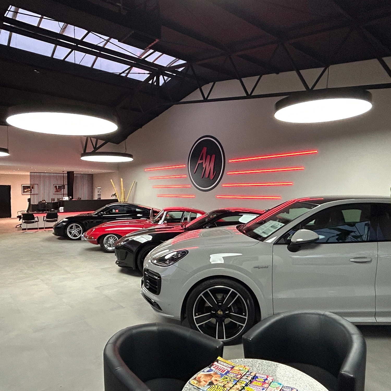 2 Auto March, Remagen Rahmen Licht translucent In einem modernen Autohaus stehen mehrere Luxusautos in Reihe. Große runde Deckenleuchten erhellen den Raum, ein Neon-AM-Logo ist an der Wand. Im Vordergrund befinden sich zwei schwarze Stühle und ein Tisch mit Zeitschriften.