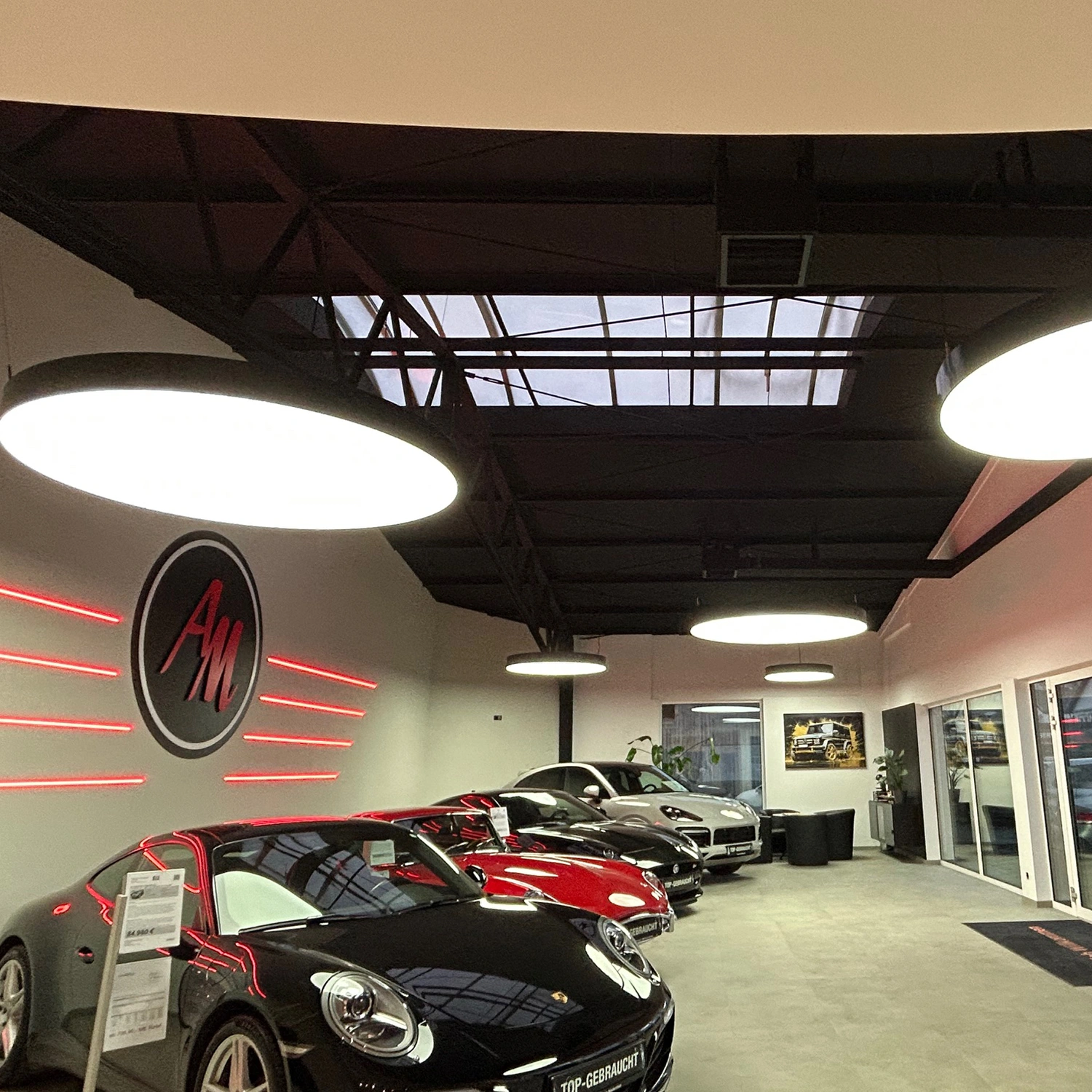 3 Auto March, Remagen Rahmen Licht translucent Moderner Autoausstellungsraum mit mehreren Sportwagen in Schwarz, Rot und Silber unter großen runden Deckenleuchten, Glasdach und rotem AM-Logo an der Wand.
