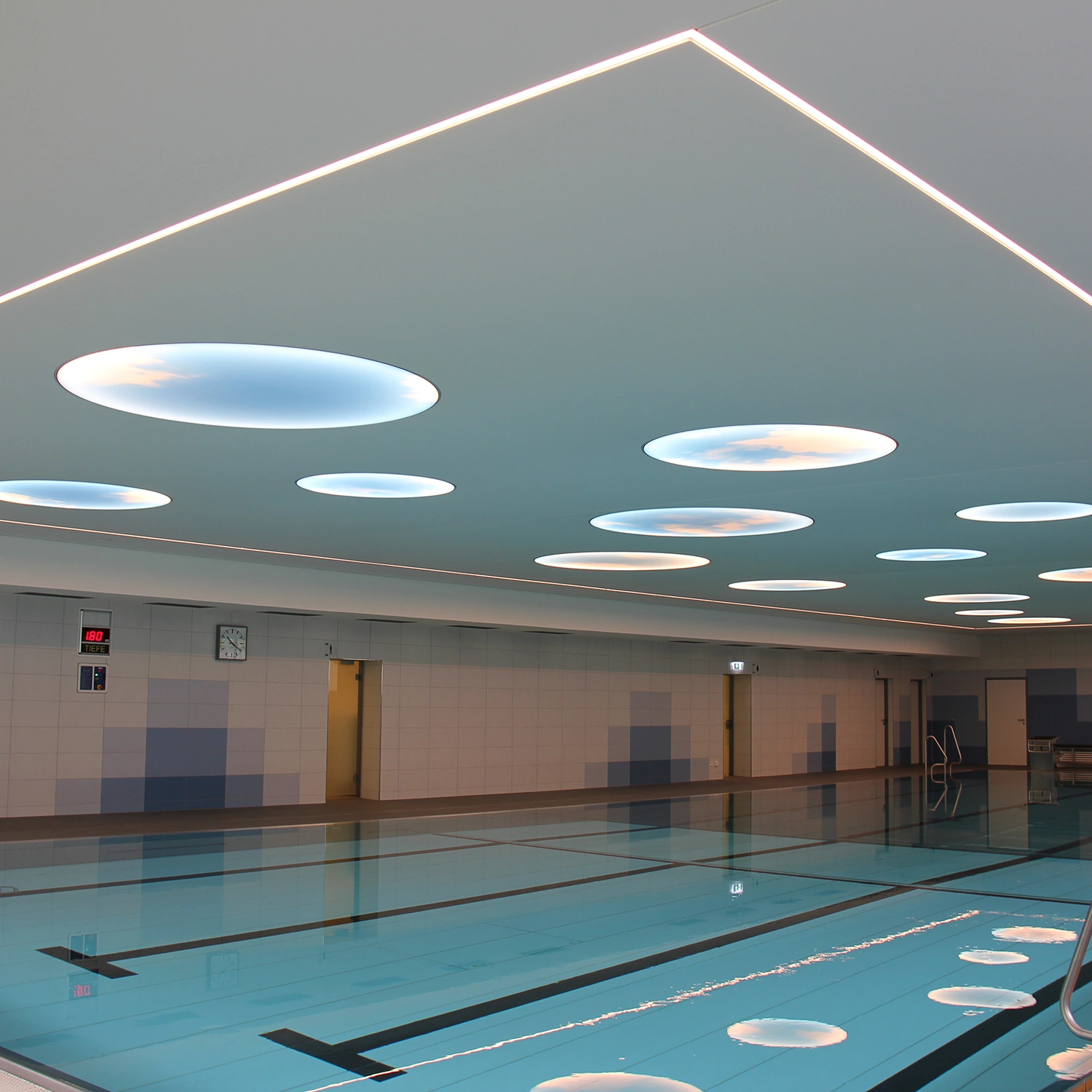 Hallenbad Erolzheim mit klarem Wasser, gefliesten Wänden und Deckenleuchten in Wolkenoptik, Türen an der Stirnseite.