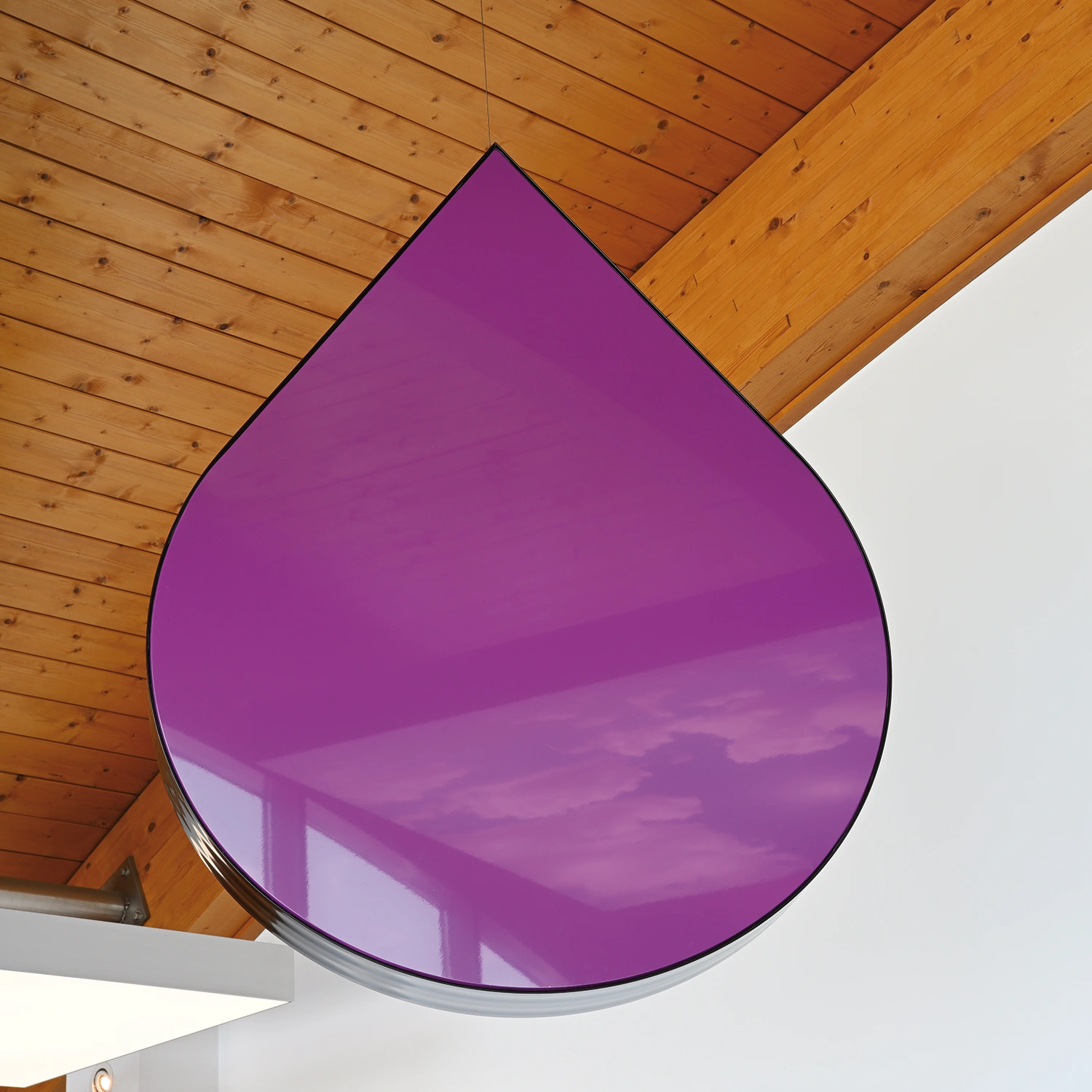 FFV_6826 Ein großes, glänzendes, violettes Objekt in Tropfenform hängt an einer Holzdecke mit sichtbaren Balken und weißen Wänden.