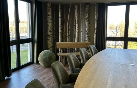 Moderner Besprechungsraum mit dunklen Stühlen um einen Holztisch, Birkenstämmen an der Wand, Blätterdecke und großen Fenstern.