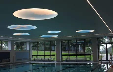 Im Hallenbad Erolzheim spiegelt sich das Wasser des Innenpools, große Fenster zeigen ins Grüne, runde Einbauleuchten an der Decke.
