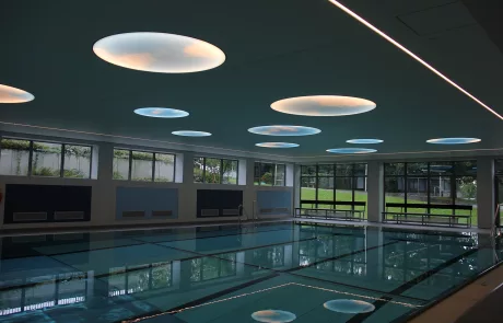 Hallenbad Erolzheim mit klarem Wasser, großen Fenstern und weißen Fliesenwänden; runde Deckenleuchten spiegeln sich im Wasser.