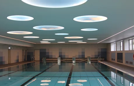 Modernes Hallenbad in Erolzheim mit Schwimmbecken, markierten Bahnen, runden Deckenleuchten, Fliesenwänden und großen Fenstern.