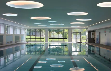 Das Erolzheim Hallenbad zeigt ein modernes Schwimmbecken, große runde Deckenleuchten, hohe Fenster und klare Bahnmarkierungen.