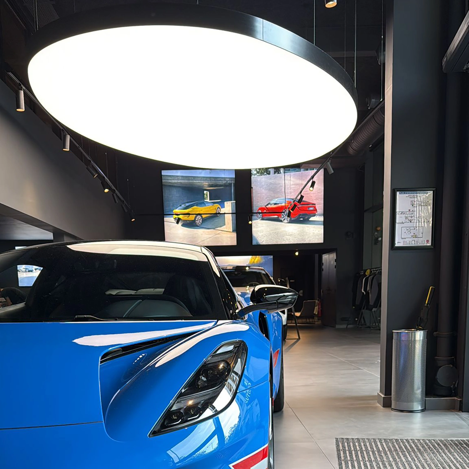 Lotus Ein blauer Sportwagen steht in einem modernen Showroom mit dunklen Wänden unter einer großen runden Deckenleuchte. Im Hintergrund sind zwei Bildschirme mit gelben und roten Sportwagen zu sehen.