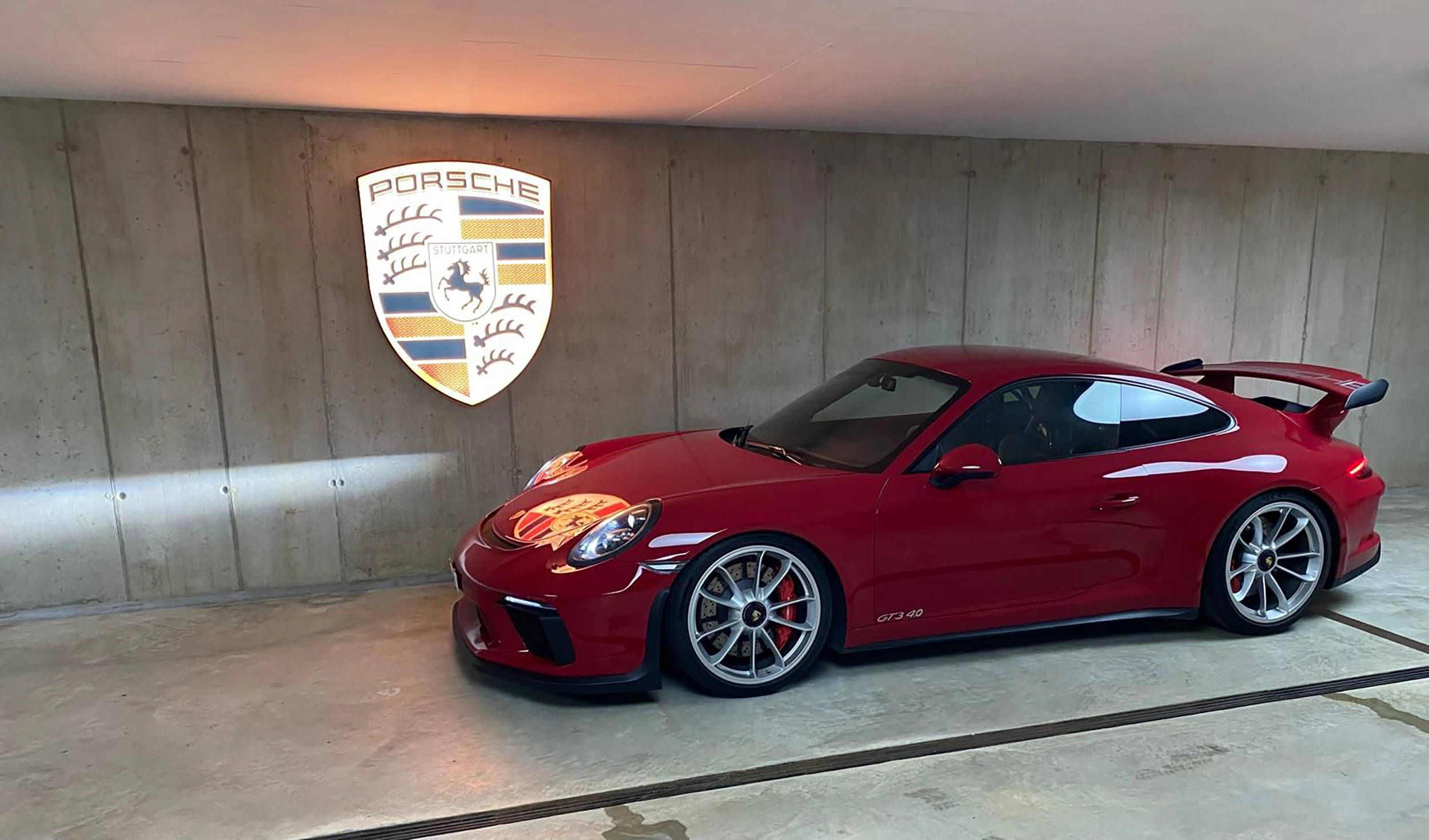 Porsche Emblem Garage2 Ein roter Porsche 911 GT3 mit silbernen Felgen und Heckspoiler steht in einer Halle vor einer Betonwand mit beleuchtetem Porsche-Logo.