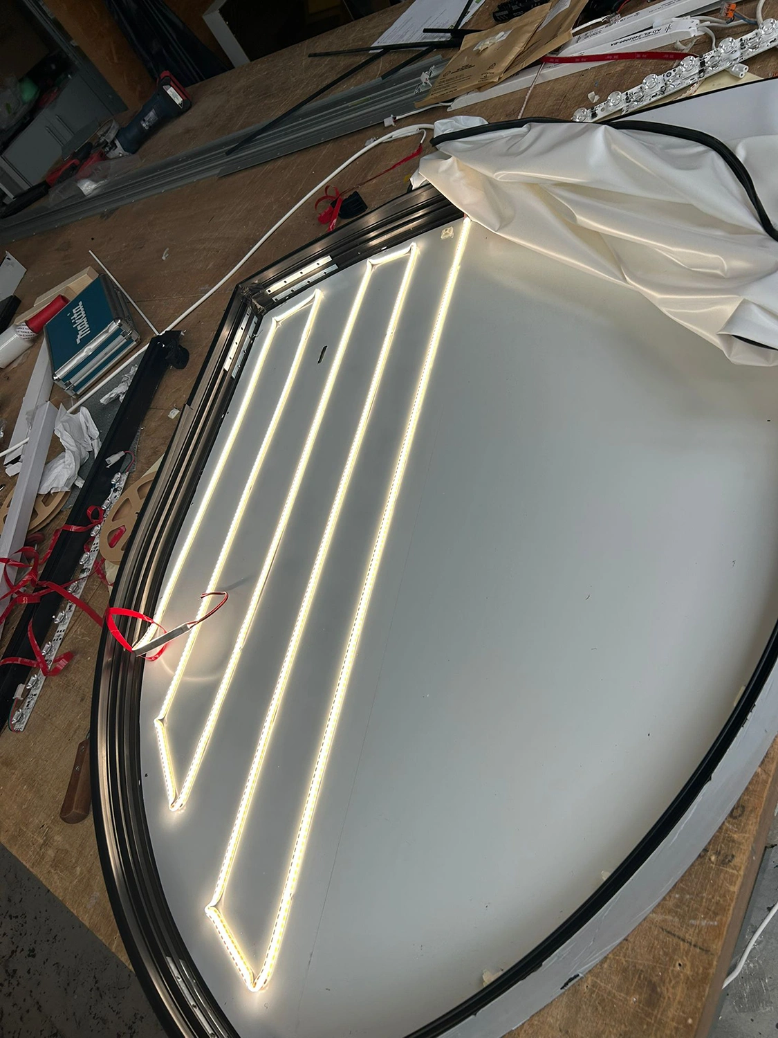 Porsche Emblem LED an Ein großer, gebogener Rahmen mit innenliegenden LED-Streifen liegt auf einer Werkbank, umgeben von Werkzeug und weißen Stoffbahnen.