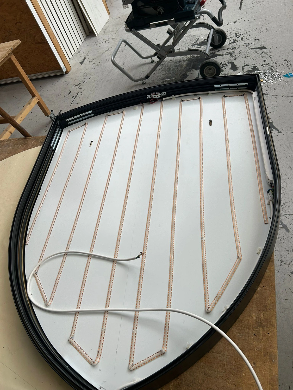 Porsche Emblem LED Ein großes, gebogenes Metallpaneel mit Zickzack-LED-Bändern und weißem Stromkabel liegt auf einer Werkstattfläche; Werkzeug im Hintergrund.