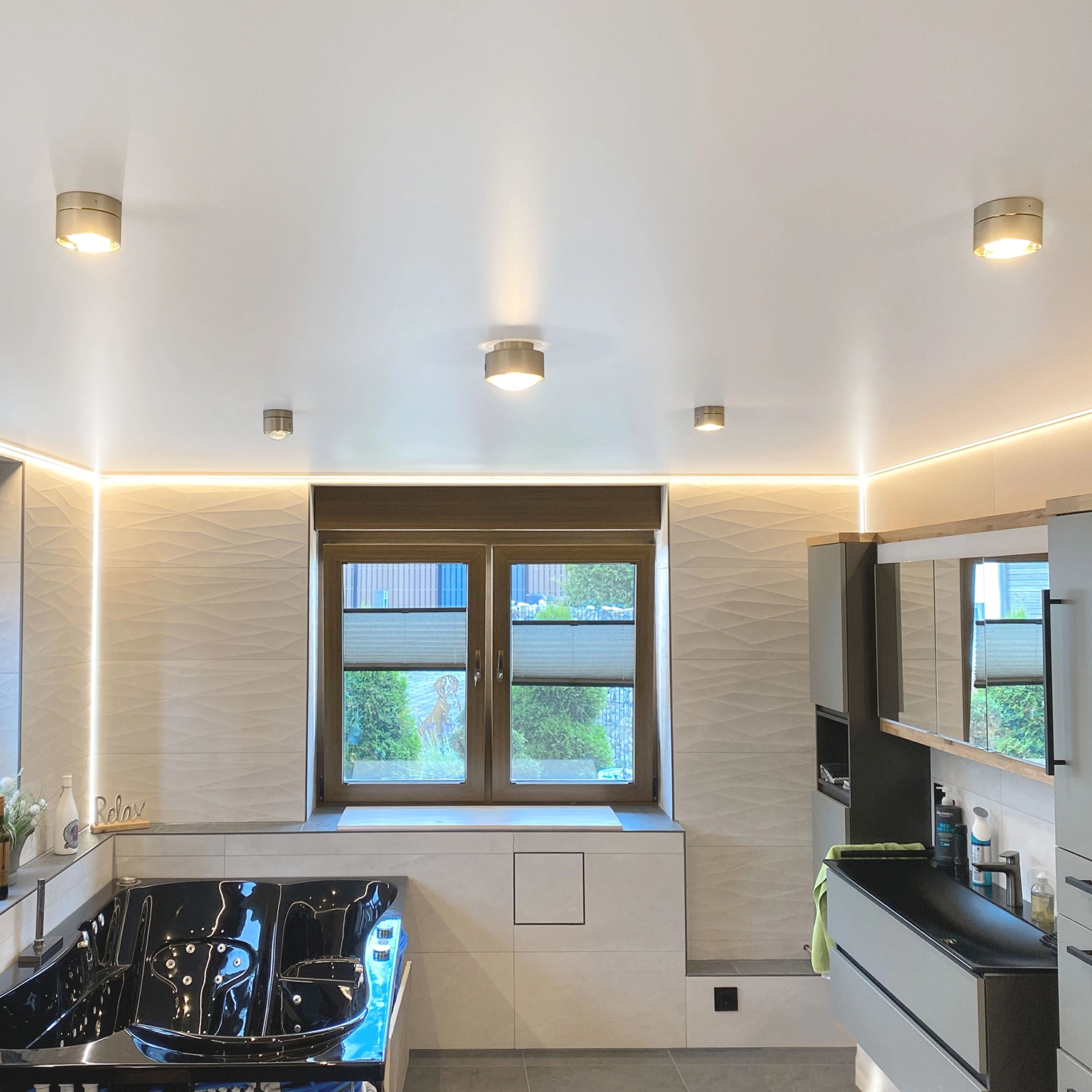 Startseite_Bad Modernes Bad mit schwarzer Badewanne, großem Fenster mit Jalousien, weißen strukturierten Wänden und Spanndecke mit Beleuchtung.