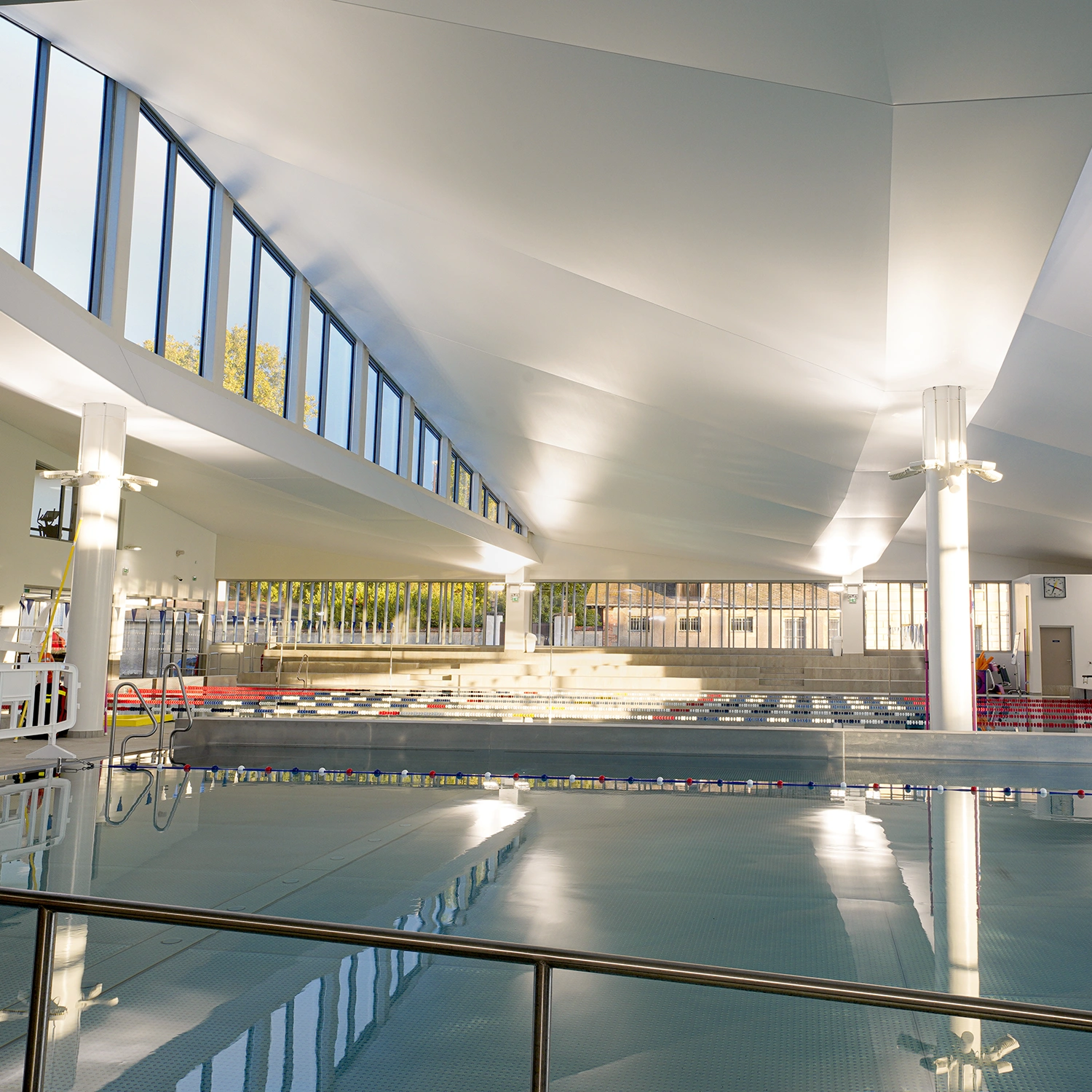 Hallenbad im Centre Aquatique de Viroflay mit klarem Wasser, großen Fenstern, Bahnteilern, Zuschauerplätzen und hoher Decke.