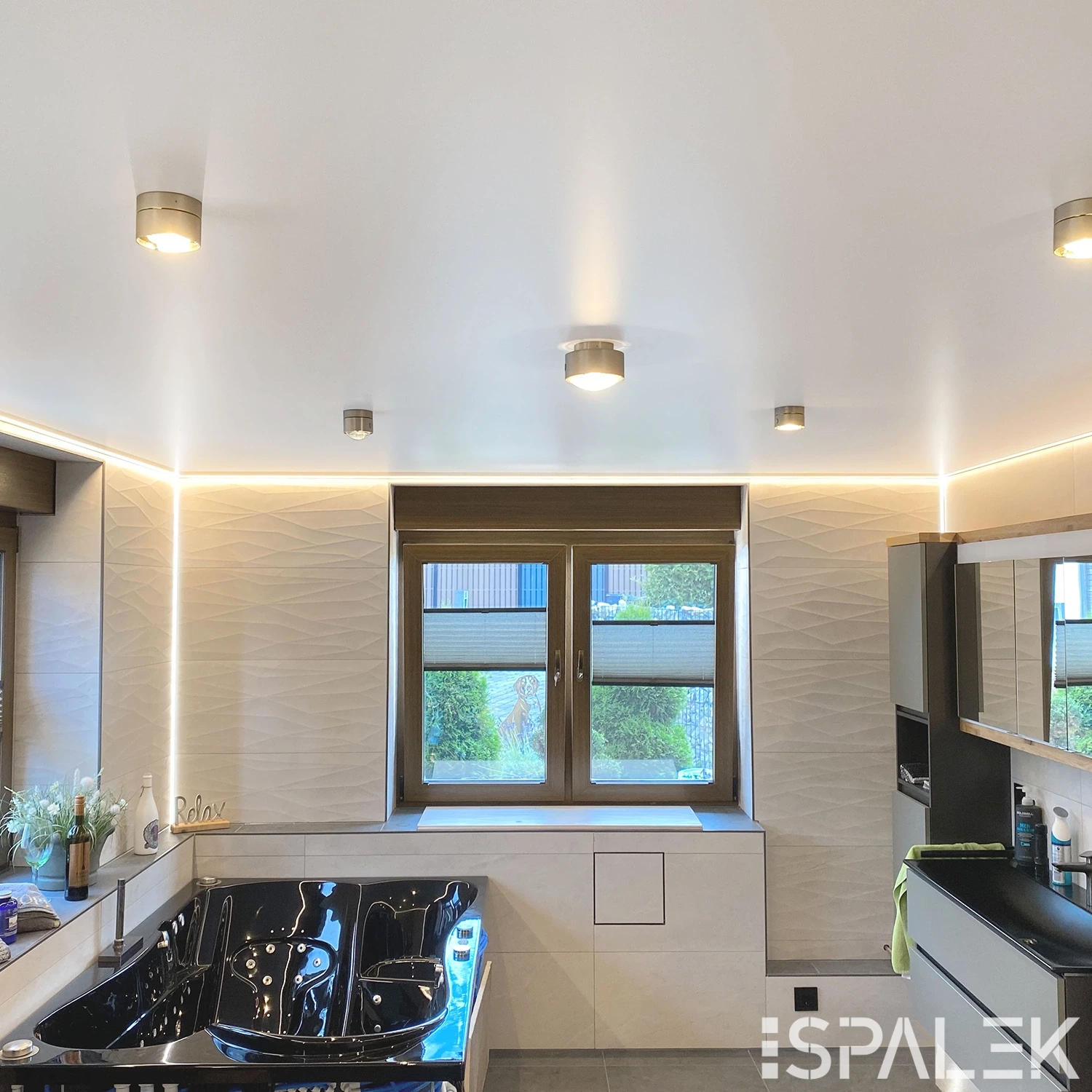 Modernes Badezimmer mit großer schwarzer Badewanne, beigen Wänden, breitem Fenster mit Jalousien, Einbauregalen und Strahlern.