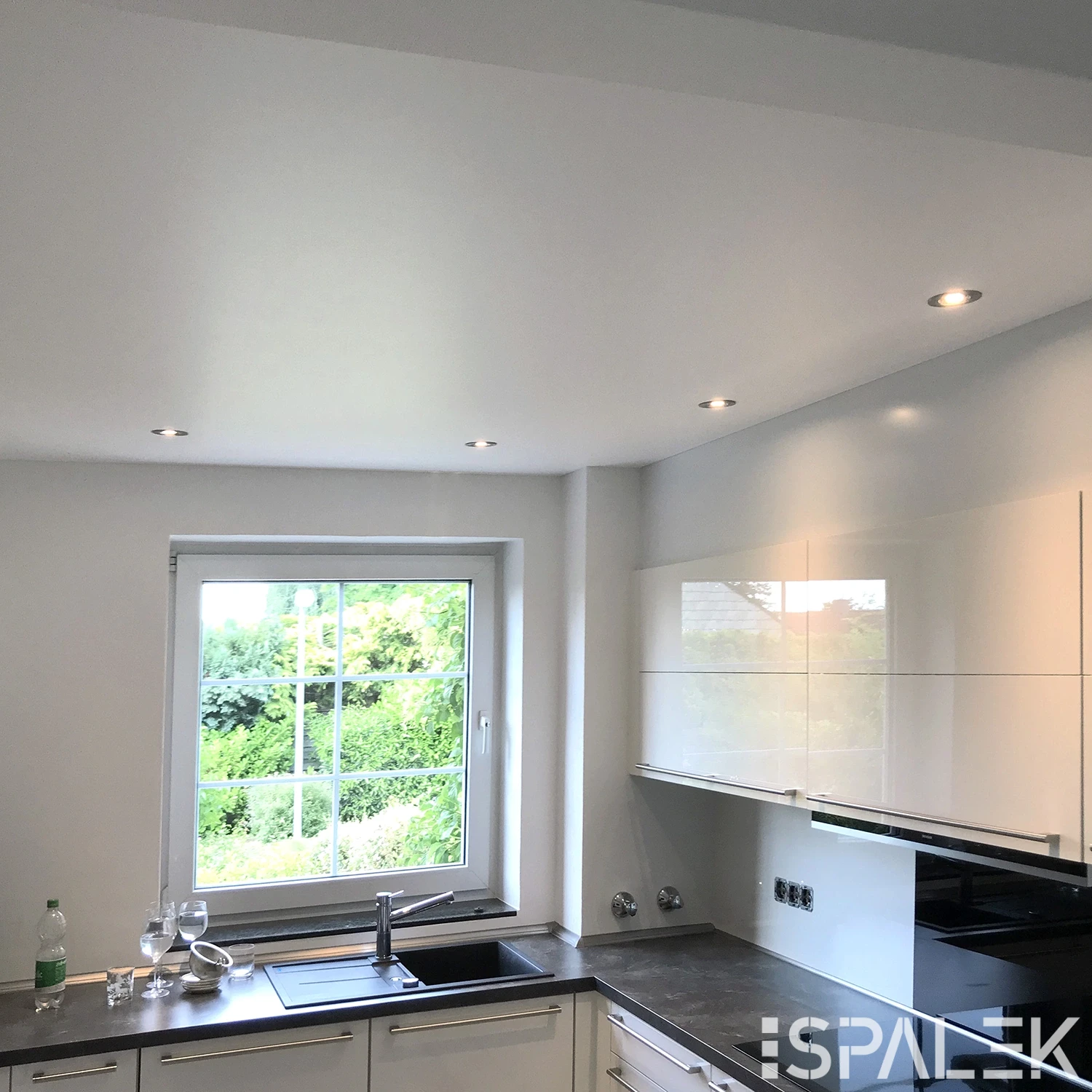 Moderne weiße Küche mit Seidenmatten-Fronten, Einbaugeräten, dunkler Arbeitsplatte und Spüle unter dem Fenster. Spots an der Decke. Zwei Gläser und eine Flasche stehen auf der Arbeitsfläche.