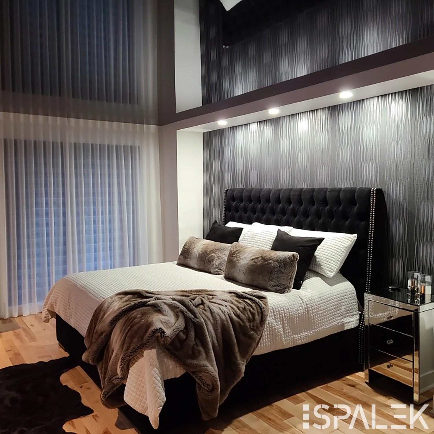 Modernes Schlafzimmer mit großem Bett, weißer Bettwäsche, schwarz-grauen Kissen, Fellplaid, Holzfußboden und Spiegelnachttisch.