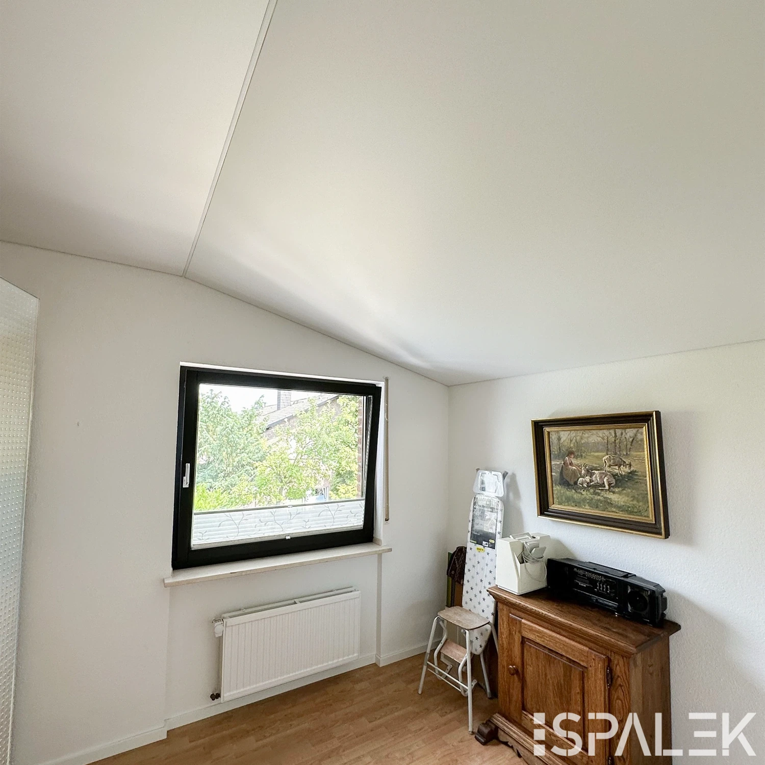 Helles Schlafzimmer mit weißen Wänden, schräger Decke, großem Fenster, Heizkörper, Holzschrank, Trittleiter und Wandbild.