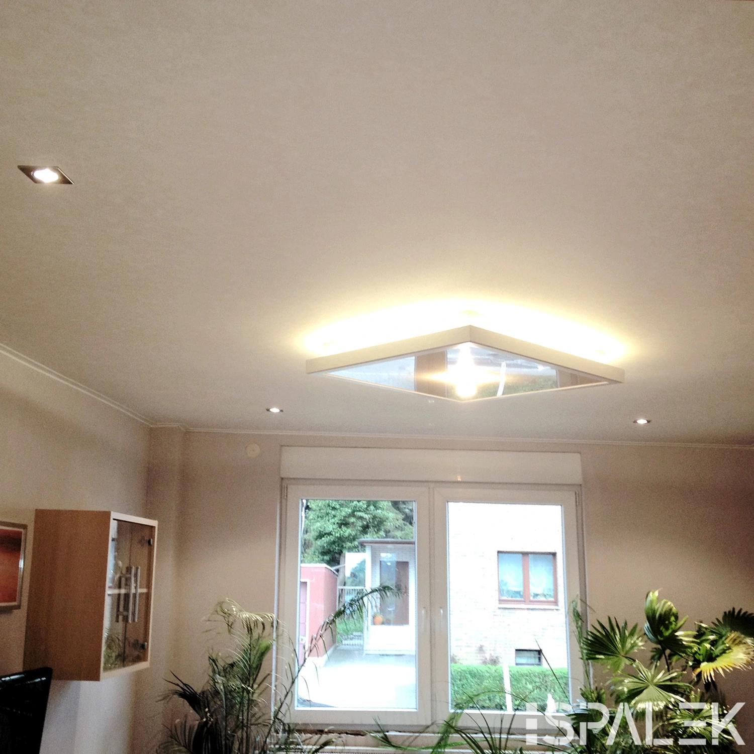 Helles, modernes Wohnzimmer mit weißer Decke, Einbaustrahlern, großem geometrischen Leuchtobjekt, Lichtleiste und Fenster zum Garten.